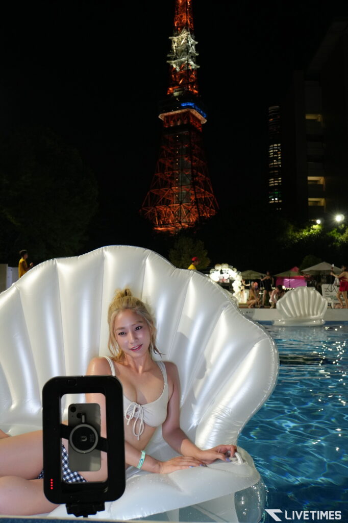 Woman in a white swimsuit sitting in a large white inflatable seashell float in a pool at night, Tokyo Tower lit red in the background, handheld phone in foreground.