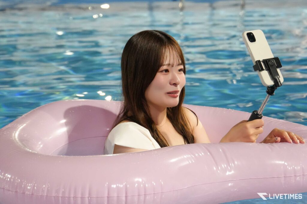 Woman in a pink inflatable pool ring taking a selfie with a white smartphone on a selfie stick, blue pool water in the background.