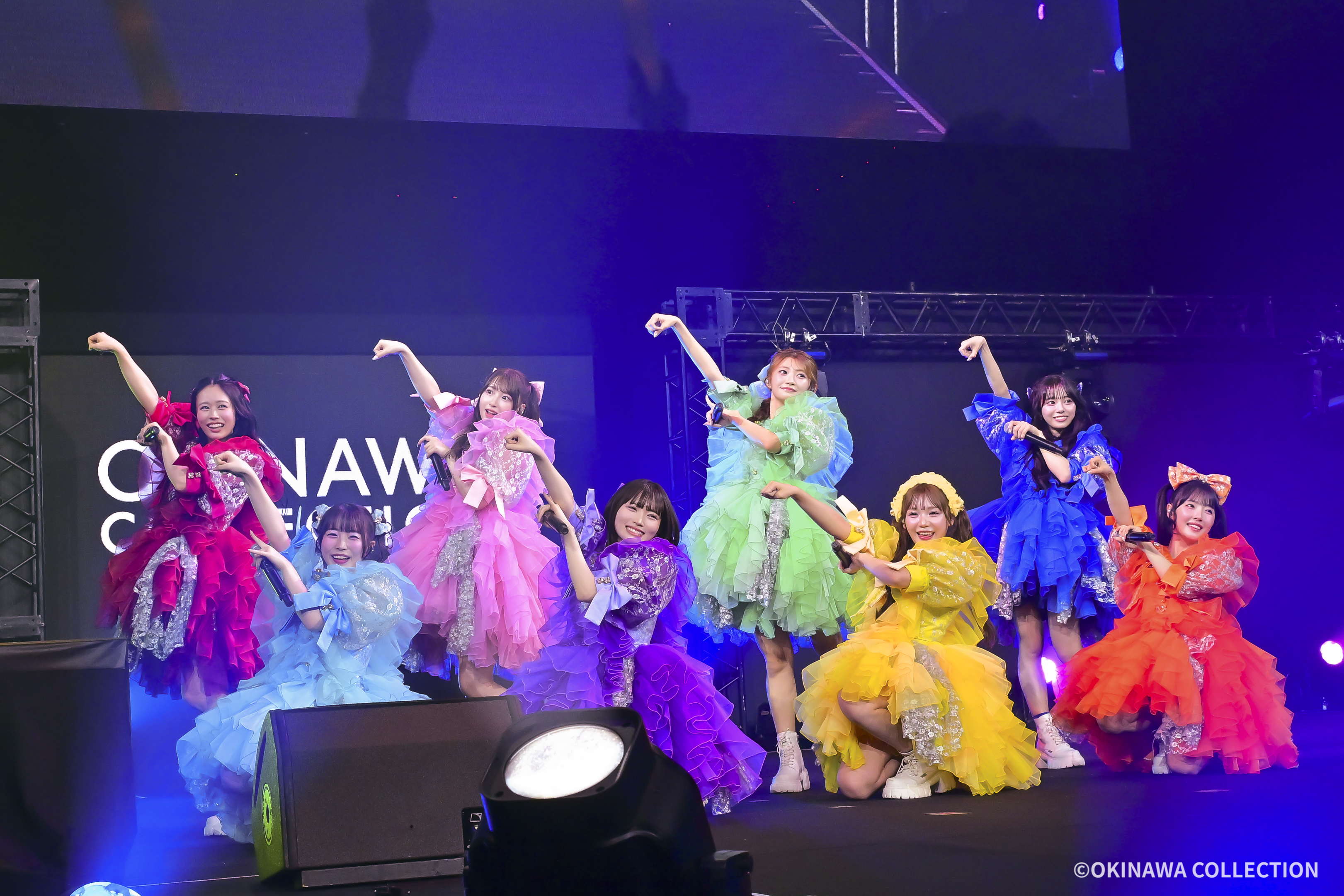 Group of performers in bright, layered dresses posing with arms raised on a stage. They wear rainbow colors (red, pink, blue, green, yellow).