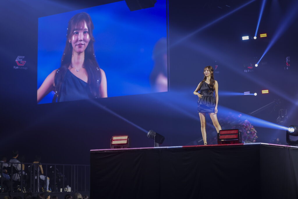 A female performer on stage in a dark blue dress, standing under bright spotlights with a large screen behind her showing a close-up image of her face.