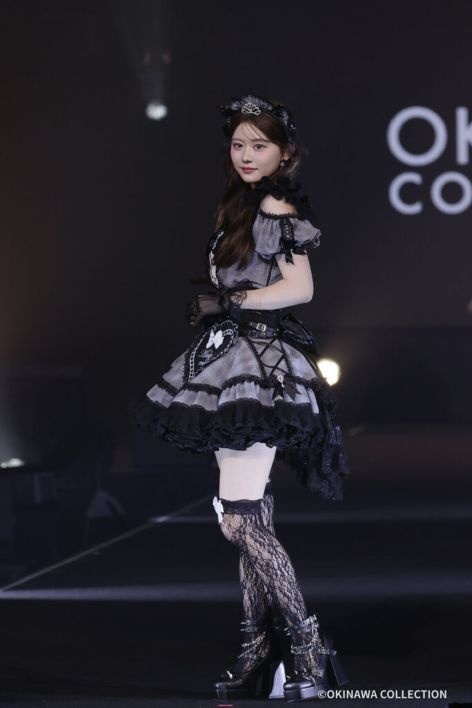 Woman in a Gothic Lolita-inspired costume on stage, wearing a gray and black layered dress with lace, gloves, and a tiara.