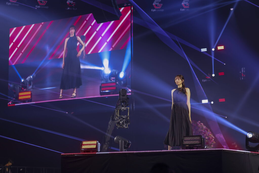Two women in evening gowns stand on a dark, neon-lit stage with a large pink LED screen behind them and camera equipment nearby.
