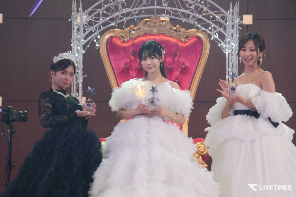 Three women in formal gowns on stage holding trophies, centered by an ornate red throne backdrop.