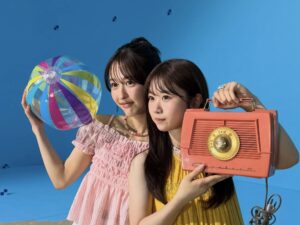 Two young women pose with a rainbow-striped beach ball and a large orange vintage radio against a blue backdrop.
