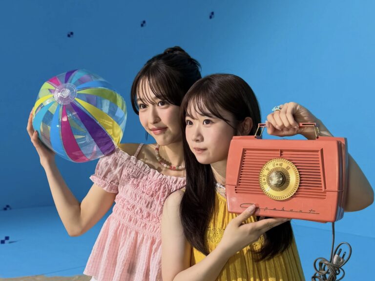 Two young women pose with a rainbow-striped beach ball and a large orange vintage radio against a blue backdrop.