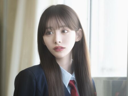 Young woman in a navy blazer and red tie, looking to the side by a bright window.