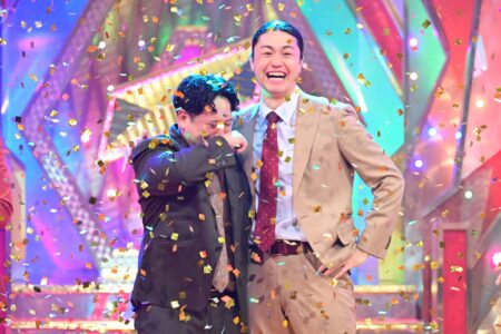 Two men in suits celebrate on a colorful stage as gold confetti rains down.