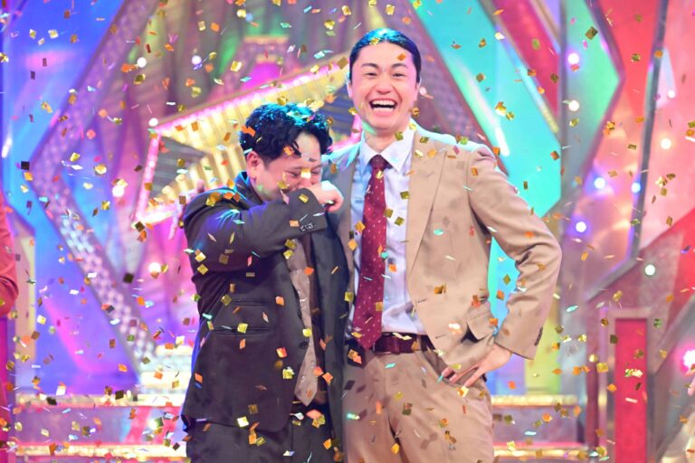 Two men in suits celebrate on a colorful stage as gold confetti rains down.