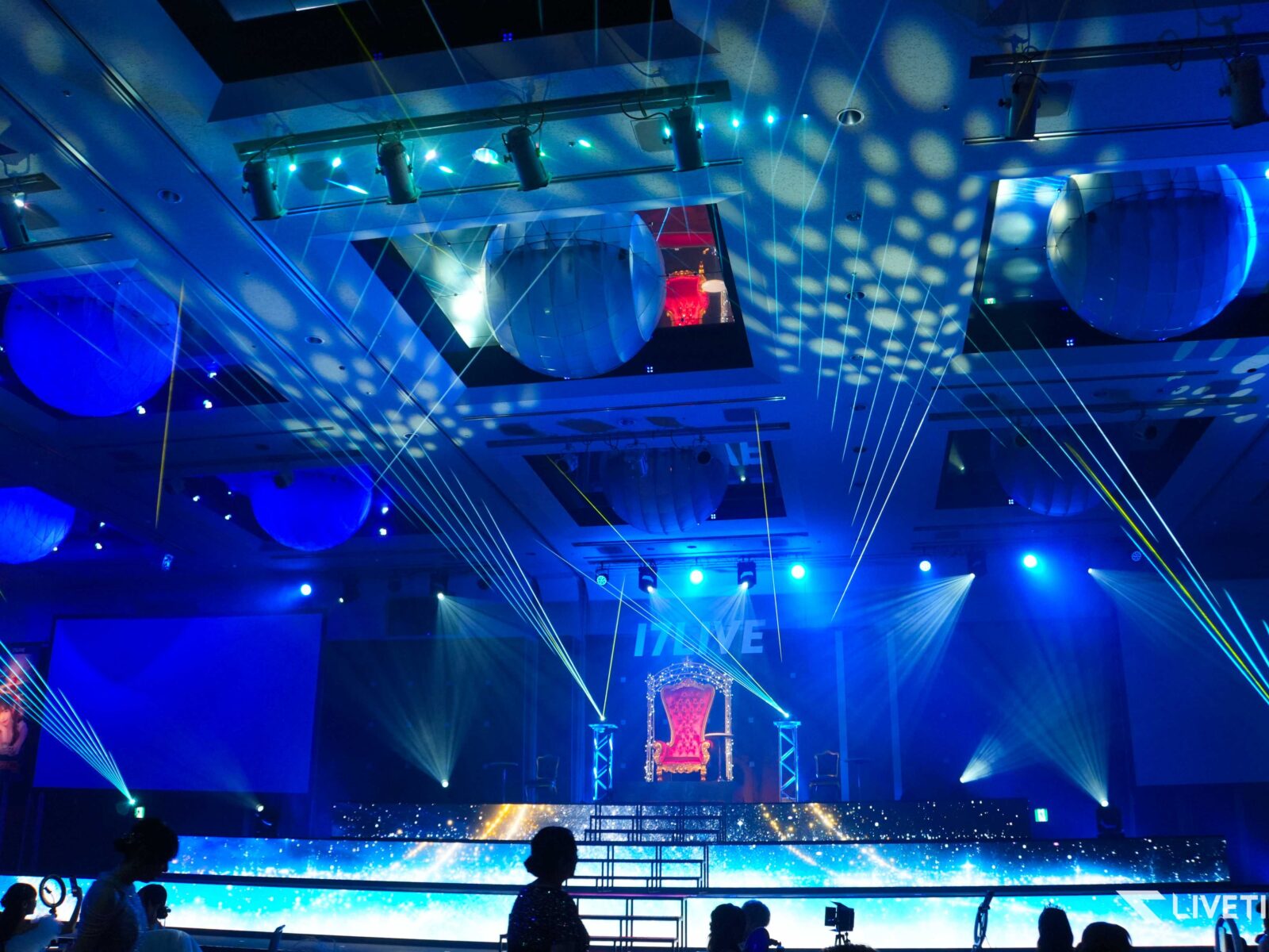Stage set with blue lighting, laser beams, and a red ornate throne at center.