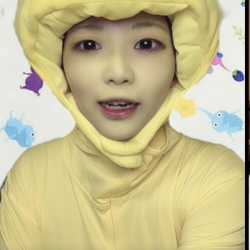 Person wearing a yellow duck-themed hooded suit, smiling at the camera, set against colorful background shapes.