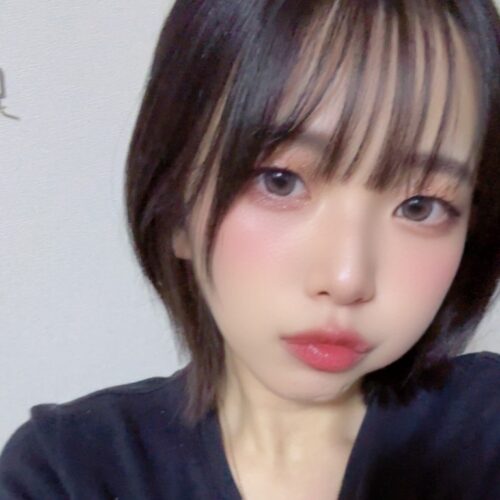 Close-up portrait of a young woman with short black hair and straight bangs, wearing a dark top and light makeup, looking at the camera against a plain wall.