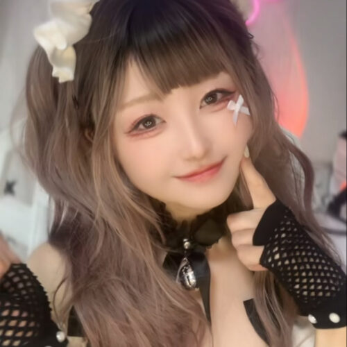 Young woman in maid cosplay taking a selfie, wearing a white frilled headband and black mesh gloves, smiling at the camera.