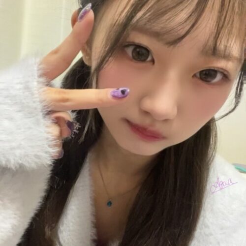 Close-up selfie of a young woman indoors, making a peace sign with her fingers, wearing a fluffy white sweater and a blue pendant necklace.