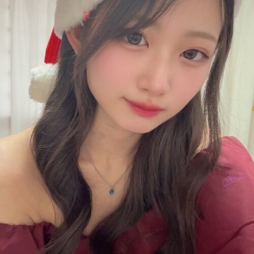 Young woman in a Santa hat and burgundy off-the-shoulder top, wearing a blue pendant necklace, looking at the camera.
