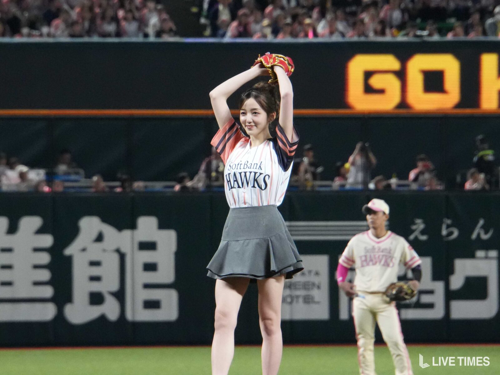 Female baseball player in a Hawks uniform raises red glove overhead on the field, smiling at the crowd.