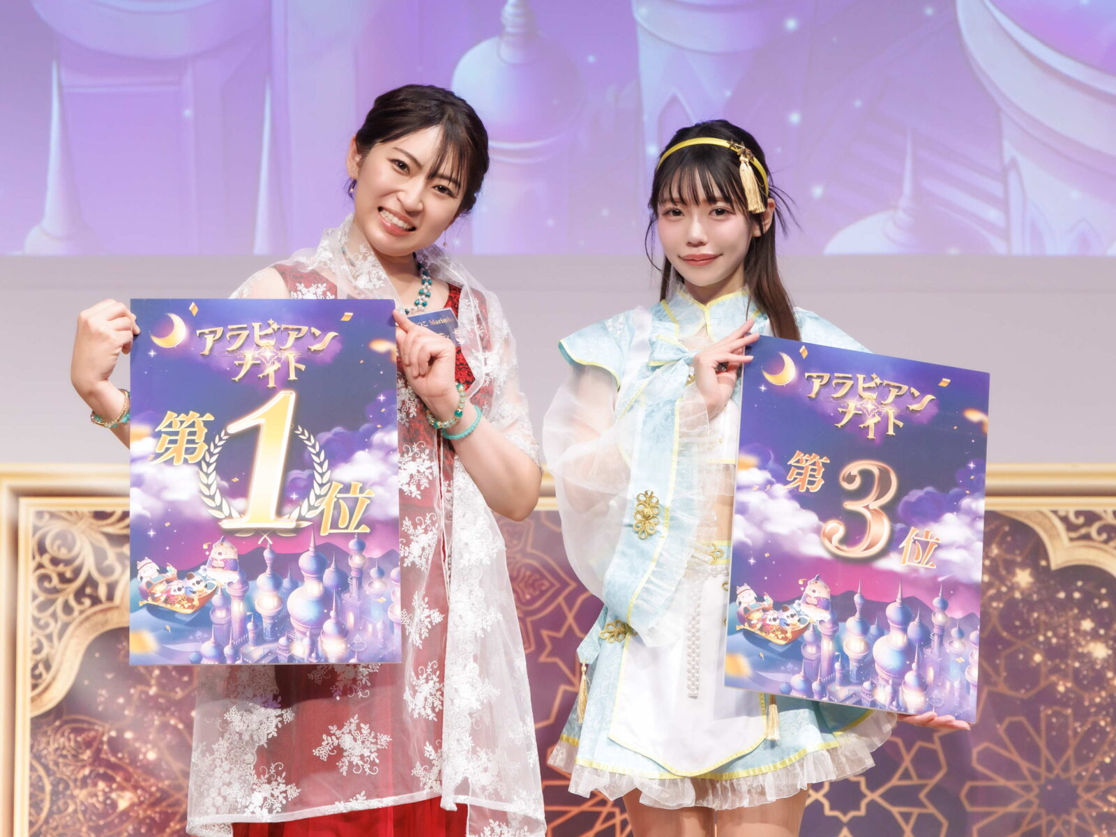 Two women in ornate costumes smile while holding large purple award posters with first place text on a stage backdrop.