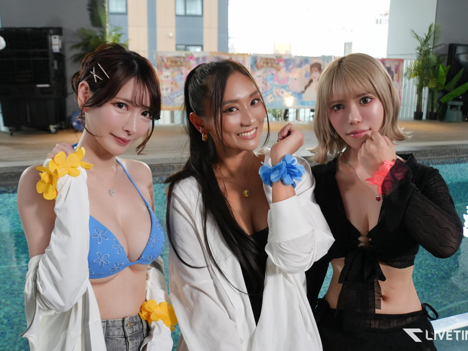 Three young women pose by a pool, wearing bikinis and flower accessories, smiling at the camera.