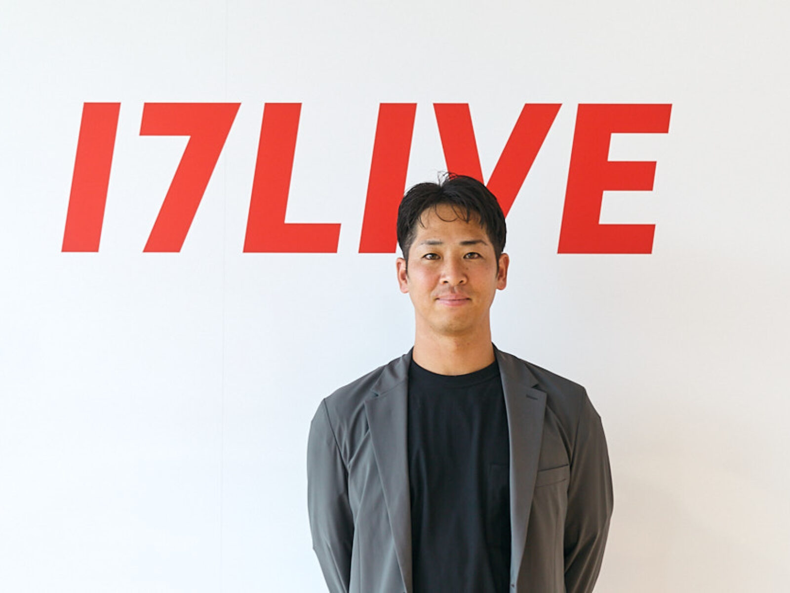 Man in a gray blazer stands before a white wall with a red 17LIVE logo behind him.